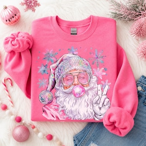 May include: A bright pink crewneck sweatshirt featuring a graphic of Santa Claus wearing a disco ball hat and glasses, blowing a pink bubblegum bubble, and making a peace sign. The design is surrounded by light blue and pink snowflakes.