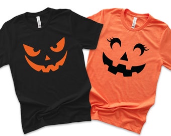 May include: Two Halloween-themed t-shirts. One black t-shirt features an orange jack-o'-lantern face with an angry expression. The other is an orange t-shirt with a smiling jack-o'-lantern face with eyelashes. Both shirts are short-sleeved.