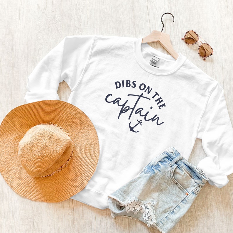 Puede incluir: Sudadera blanca con el texto "Dibs on the captain" y un gr&aacute;fico de ancla. La sudadera est&aacute; en una percha con un sombrero de paja y gafas de sol.