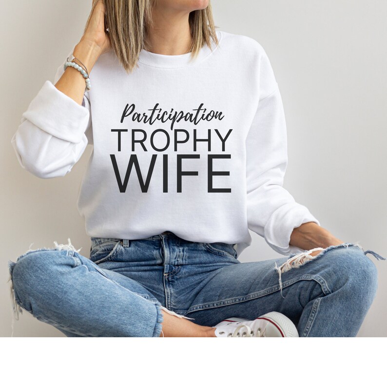 K&ouml;nnte beinhalten: Wei&szlig;er Pullover mit dem Text "Participation Trophy Wife" in schwarzen Buchstaben.