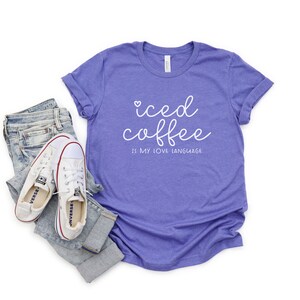 Puede incluir: Una camiseta morada con el texto "iced coffee is my love language" en letras blancas. La camiseta se muestra sobre un fondo blanco con un par de zapatillas blancas y unos vaqueros azules.