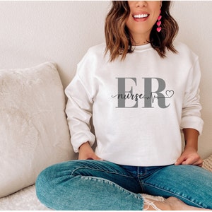 Puede incluir: Una sudadera blanca con el texto "ER nurse" en gris, una l&iacute;nea de latidos y un s&iacute;mbolo de coraz&oacute;n. La sudadera es de cuello redondo y est&aacute; hecha de un material suave.
