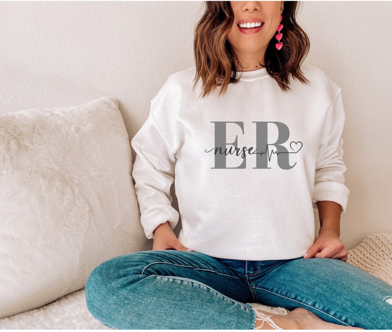 Puede incluir: Una sudadera blanca con el texto "ER nurse" en gris, una l&iacute;nea de latidos y un s&iacute;mbolo de coraz&oacute;n. La sudadera es de cuello redondo y est&aacute; hecha de un material suave.