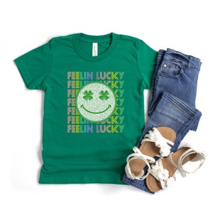 Puede incluir: Camiseta verde con un gr&aacute;fico de arco&iacute;ris "Feelin Lucky" que presenta una cara sonriente con tr&eacute;boles de cuatro hojas como ojos.