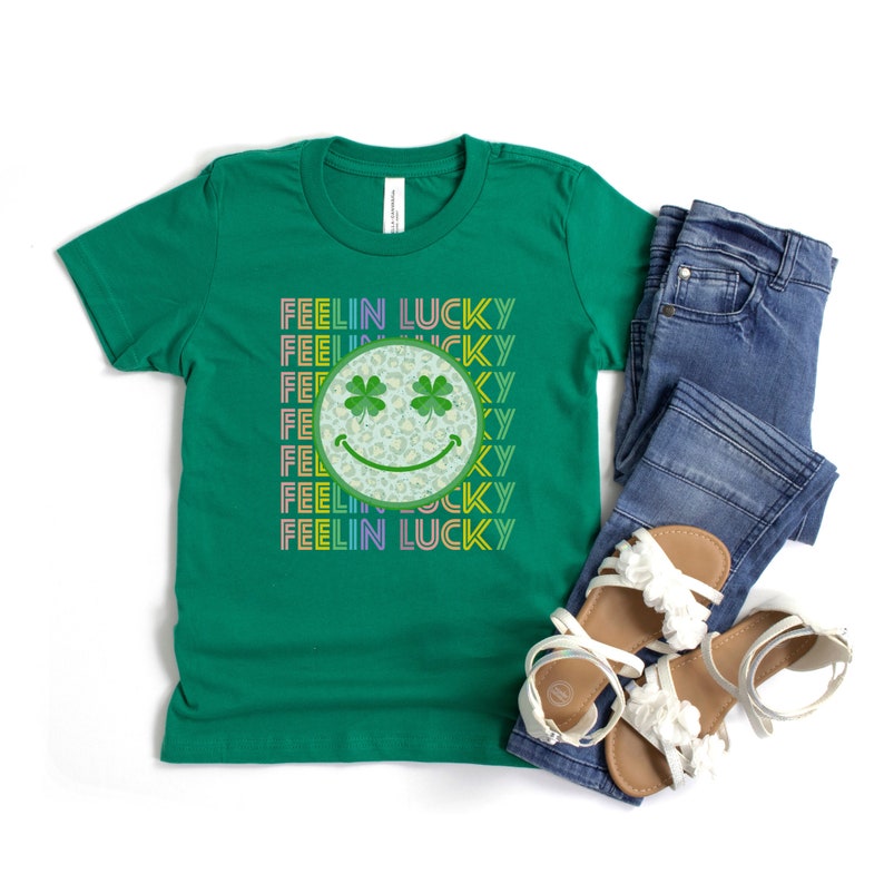 Puede incluir: Camiseta verde con un gr&aacute;fico de arco&iacute;ris "Feelin Lucky" que presenta una cara sonriente con tr&eacute;boles de cuatro hojas como ojos.