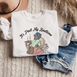 Pu&ograve; includere: Felpa bianca con una grafica di un orso che indossa un berretto da notte blu e siede su una sedia con un libro e delle candele. Il testo "It's Past My Bedtime" &egrave; stampato in nero sulla felpa.