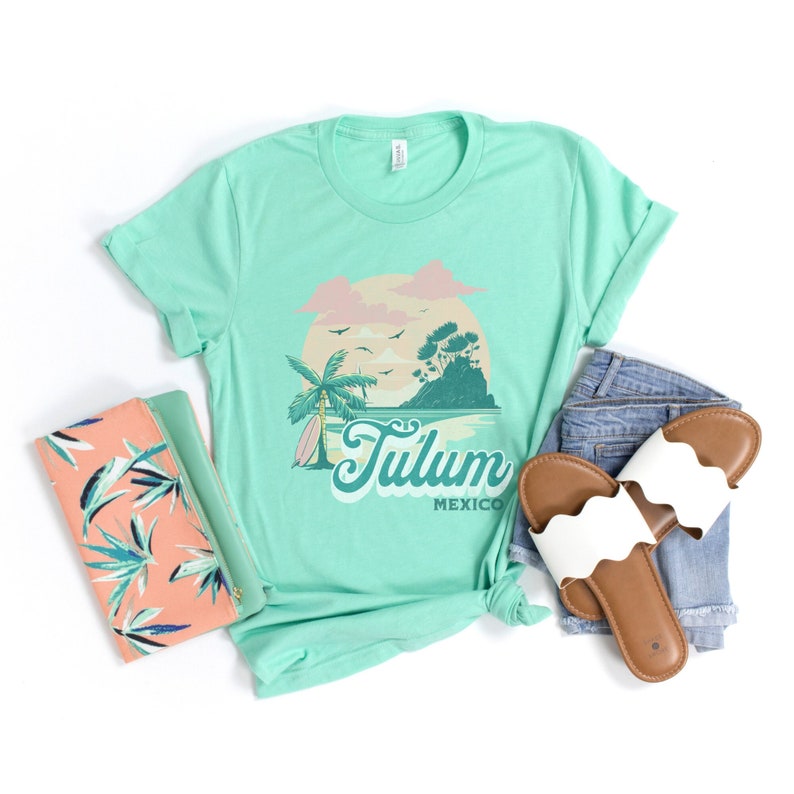 May include: A light green t-shirt with a retro graphic design of a beach scene with a palm tree, a surfboard, and the text "Tulum Mexico". The t-shirt is laid out on a white surface with a pair of white sandals and a pink and green patterned clutch.