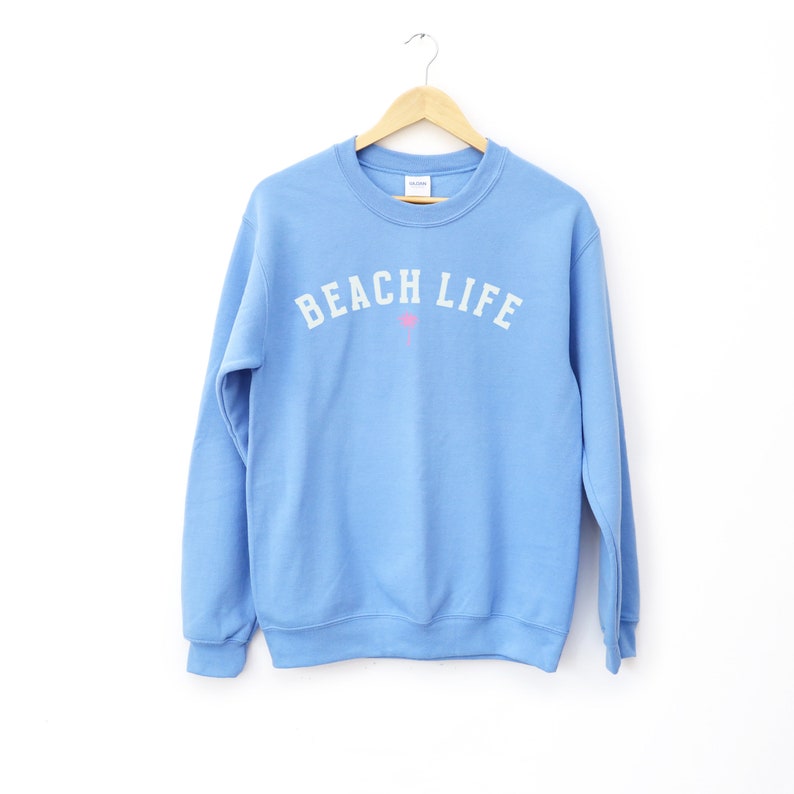 Pode incluir: Uma camisola azul claro com o texto "BEACH LIFE" em letras brancas e um &iacute;cone de palmeira rosa.