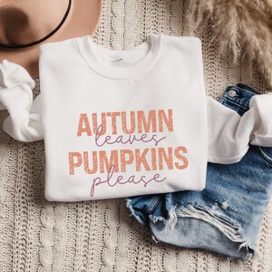 以下が含まれることがあります： オレンジと紫色の「AUTUMN leaves PUMPKINS please」の文字が入った白いスウェットシャツ。クリーム色のニットブランケットの上に折りたたまれ、デニムショーツとベージュの帽子が背景にあります。