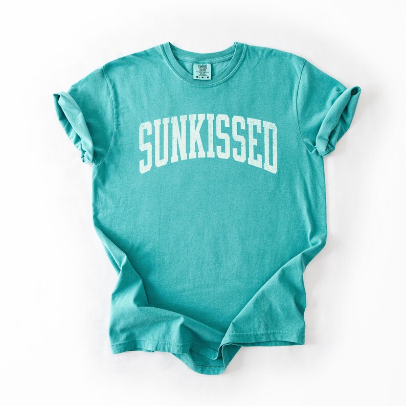 May include: A turquoise t-shirt with the word "SUNKISSED" printed in white, arched lettering. The shirt has short sleeves and a crew neck. The fabric is slightly wrinkled, and the shirt is laid flat on a white surface.