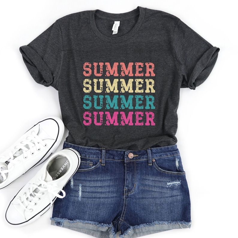 May include: Dark gray t-shirt with the word "SUMMER" in a stacked design, using retro-style lettering in shades of pink, teal, yellow, and orange. The shirt is paired with denim shorts and white canvas sneakers, creating a casual summer outfit.