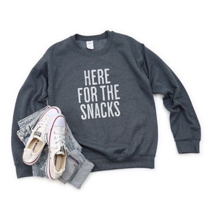May include: A dark gray sweatshirt with the text "HERE FOR THE SNACKS" printed in white on the front. The sweatshirt is laying flat on a white background with a pair of white sneakers and blue jeans.