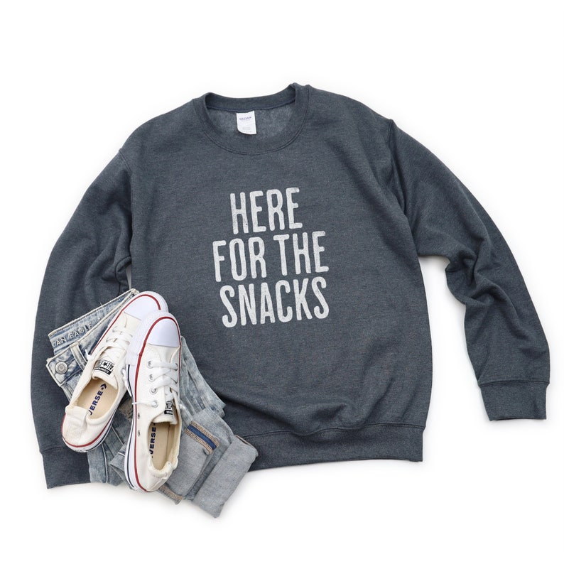 May include: A dark gray sweatshirt with the text "HERE FOR THE SNACKS" printed in white on the front. The sweatshirt is laying flat on a white background with a pair of white sneakers and blue jeans.