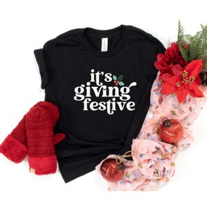 May include: Black crew neck t-shirt with the text "it's giving festive" in white. The shirt is styled with red mittens, a festive scarf, and Christmas ornaments. The design includes holly and berry accents, perfect for the holiday season.