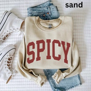 Może przedstawiać: Beżowa bluza z nadrukiem "SPICY" w kolorze bordowym z przodu. Bluza leży na białym tle z parą białych but&oacute;w sportowych i niebieskimi dżinsami.