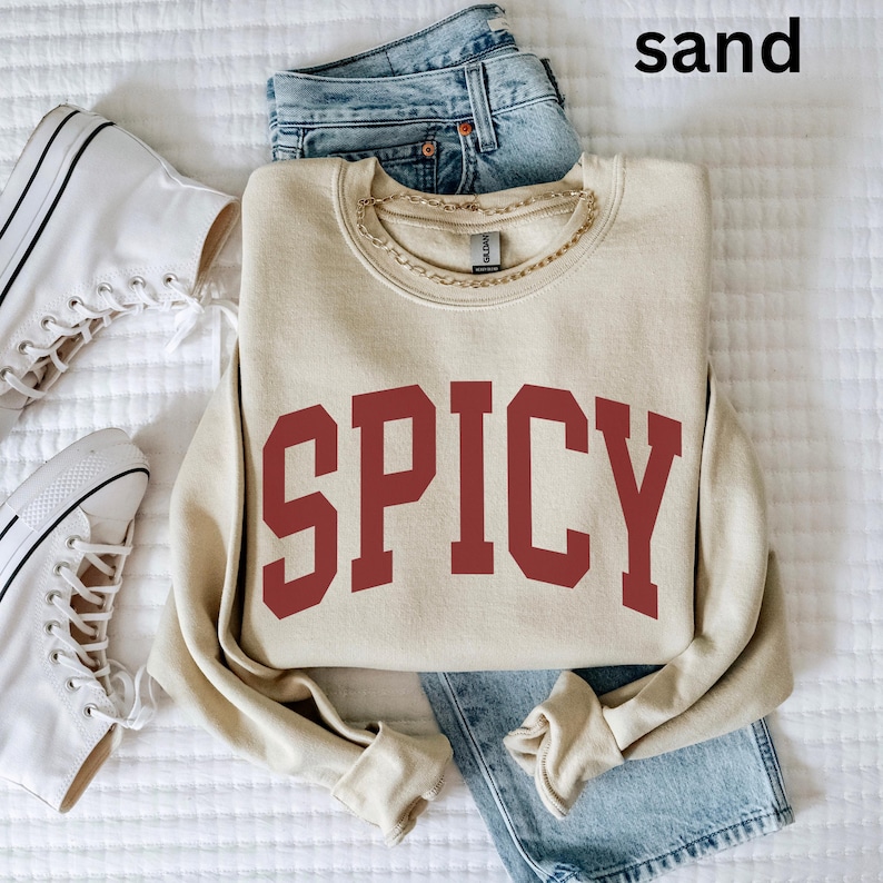 Może przedstawiać: Beżowa bluza z nadrukiem "SPICY" w kolorze bordowym z przodu. Bluza leży na białym tle z parą białych but&oacute;w sportowych i niebieskimi dżinsami.