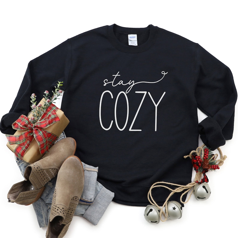 May include: Black sweatshirt with white text that reads "stay cozy" on a white background. The sweatshirt is surrounded by a variety of festive decorations, including a wrapped gift, boots, and a sprig of holly.