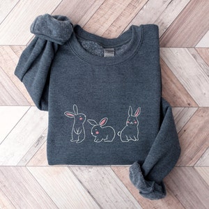 May include: A dark grey sweatshirt featuring a white outline of three bunnies. The bunnies have pink noses and ears.