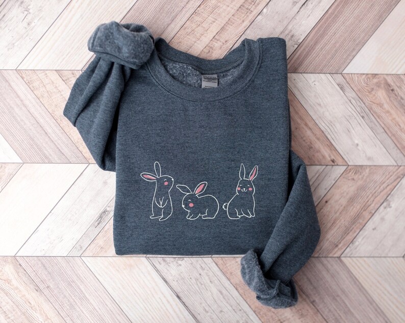 May include: A dark grey sweatshirt featuring a white outline of three bunnies. The bunnies have pink noses and ears.