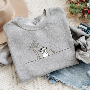 Op de afbeelding: Een grijze sweatshirt met een winterthema-illustratie. Het ontwerp toont een sneeuwpop met een hoed en sjaal, een kale boom en een hond. De illustratie is in zwart-wit met gele en lichtblauwe accenten. Een comfortabele, casual top.