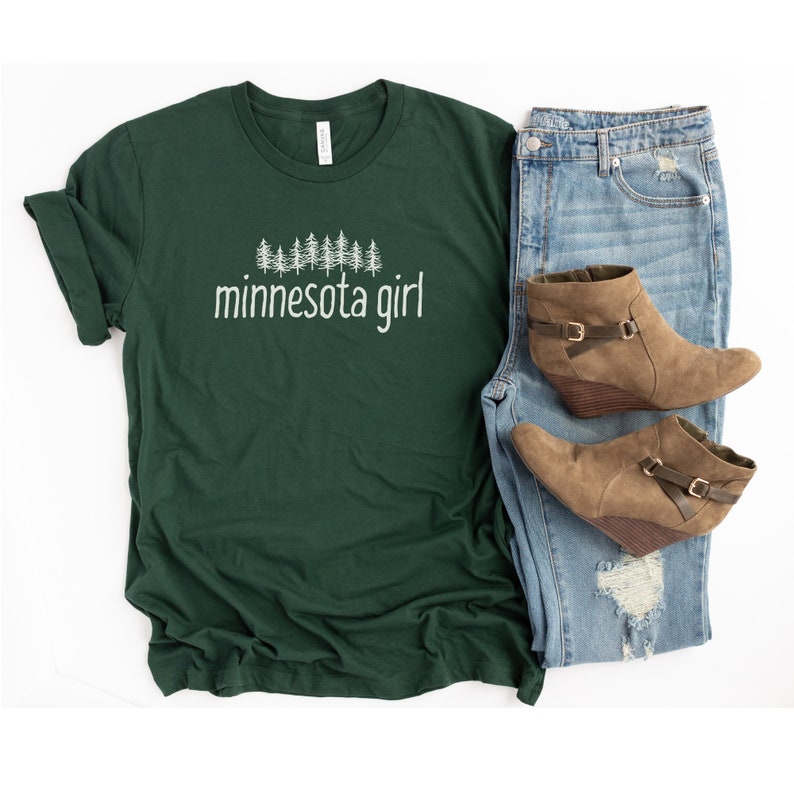 May include: A dark green t-shirt with a white graphic of pine trees and the text "minnesota girl". The t-shirt is paired with blue jeans and brown ankle boots.