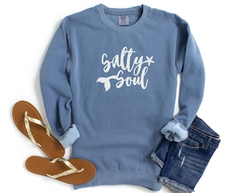 Könnte beinhalten: Ein hellblauer Pullover mit dem Schriftzug "Salty* Soul" in weißen Buchstaben. Unter dem Text befindet sich ein weißer Meerjungfrauen-Schwanz. Der Pullover liegt flach auf einem weißen Hintergrund mit einem Paar braunen Sandalen und einer blauen Jeans-Shorts.