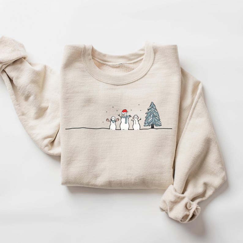 Op de afbeelding: Een cr&egrave;mekleurige sweatshirt met een minimalistisch winterontwerp. De afbeelding toont drie sneeuwpoppen met een kerstboom en vallende sneeuw. De sneeuwpoppen dragen een rode hoed en een blauwe sjaal. De sweatshirt heeft lange mouwen en een ronde hals.