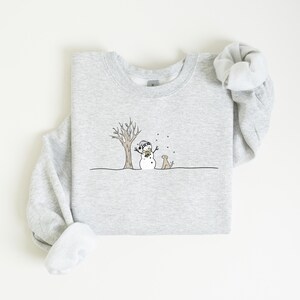 Op de afbeelding: Een lichtgrijze sweatshirt met een grillige wintersc&egrave;ne. Een sneeuwpop met een hoed en sjaal staat bij een kale boom, met een hond die toekijkt. Het ontwerp is in zwart en bruin, met vallende sneeuw.