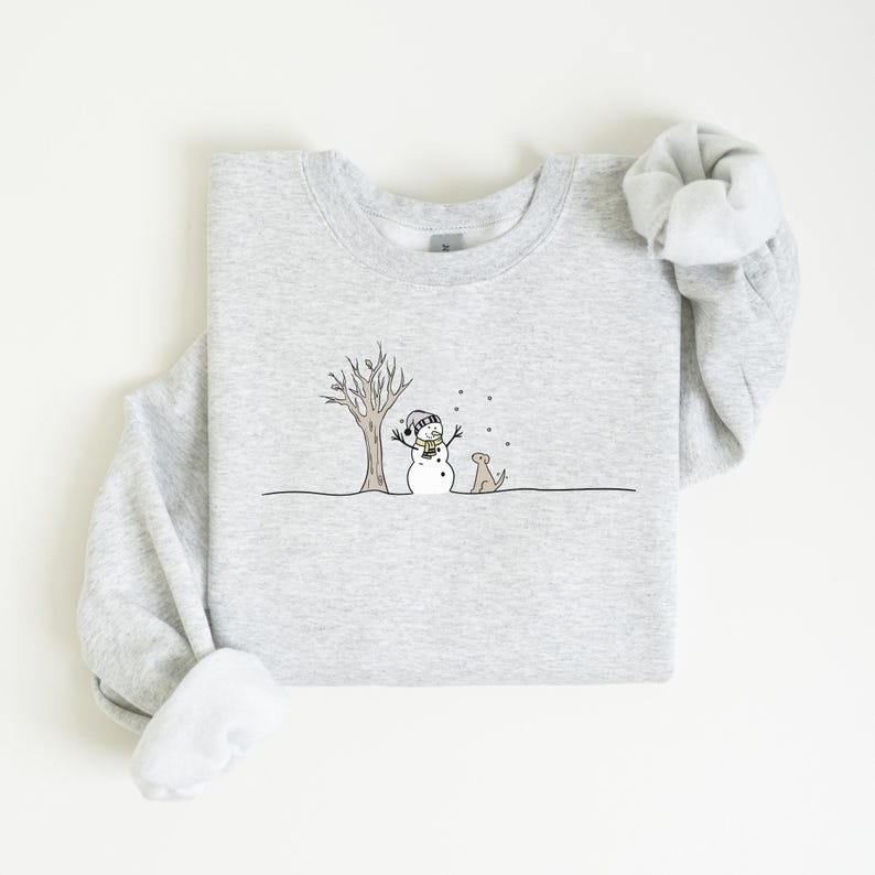 Op de afbeelding: Een lichtgrijze sweatshirt met een grillige wintersc&egrave;ne. Een sneeuwpop met een hoed en sjaal staat bij een kale boom, met een hond die toekijkt. Het ontwerp is in zwart en bruin, met vallende sneeuw.