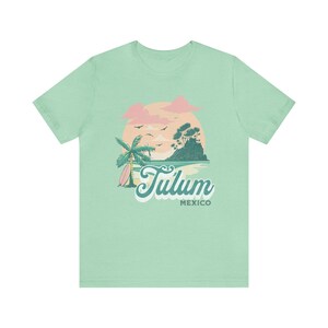 May include: A light green t-shirt with a graphic design of a palm tree, a surfboard, and a sunset over a small island. The text "Tulum Mexico" is printed on the shirt.