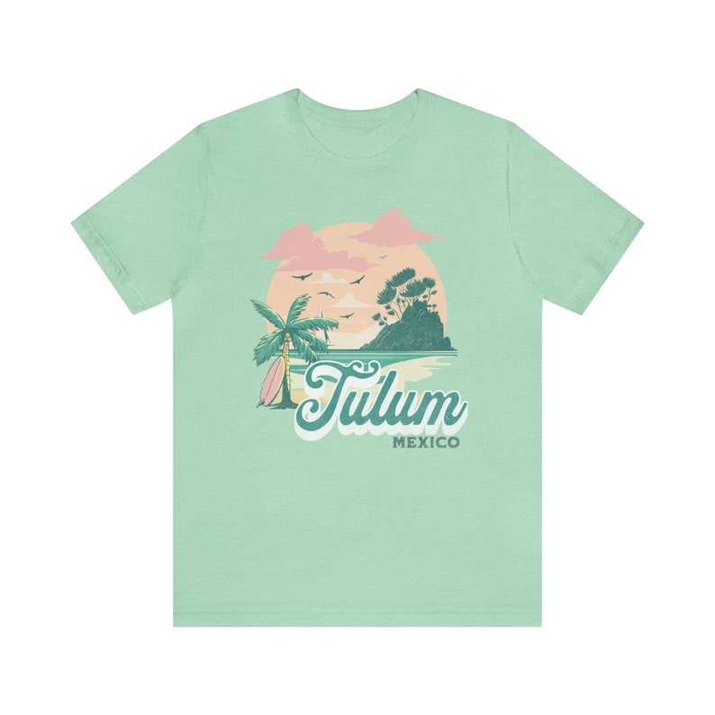 May include: A light green t-shirt with a graphic design of a palm tree, a surfboard, and a sunset over a small island. The text "Tulum Mexico" is printed on the shirt.