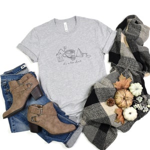 May include: A heather grey t-shirt with a graphic of tea bags, a teacup, and the text "it's a tea shirt". The shirt is styled with blue jeans, brown ankle boots, and a black and white plaid scarf.