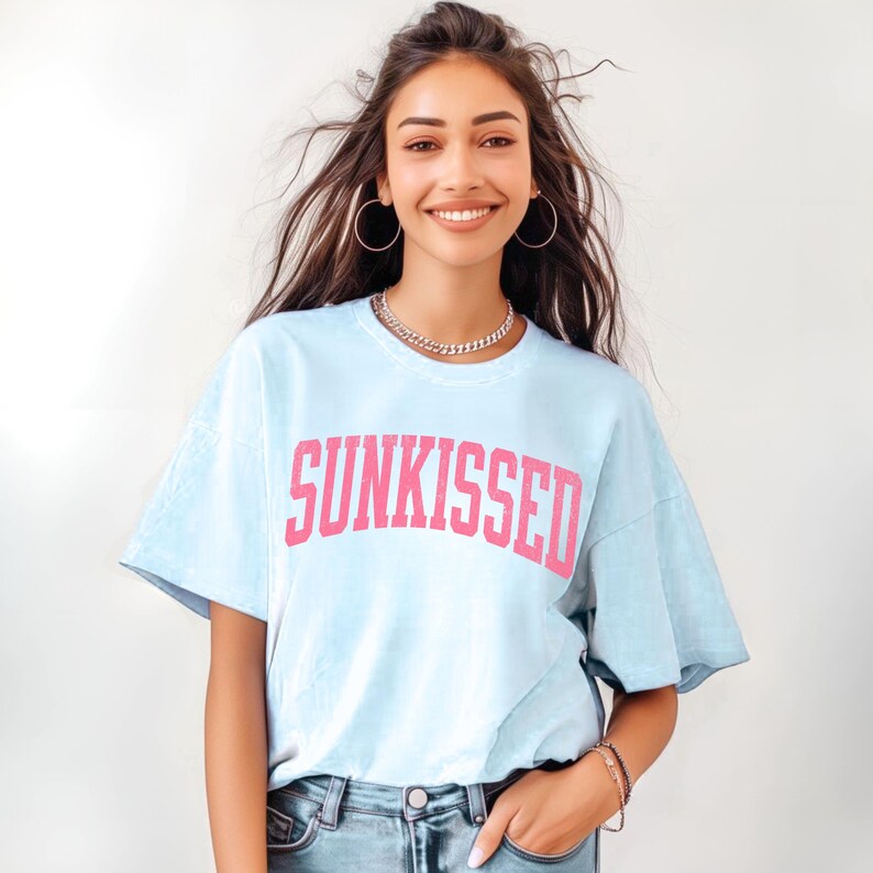 May include: Light blue t-shirt with the word "SUNKISSED" in pink block letters. The shirt has short sleeves and a relaxed fit. The model is wearing a silver chain necklace and hoop earrings.