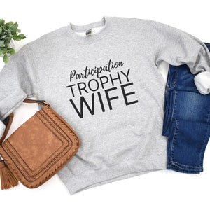 K&ouml;nnte beinhalten: Grauer Pullover mit dem Text "Participation Trophy Wife" in schwarzen Buchstaben.