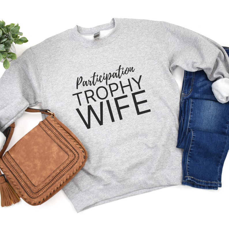 K&ouml;nnte beinhalten: Grauer Pullover mit dem Text "Participation Trophy Wife" in schwarzen Buchstaben.