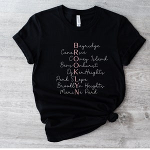 May include: Black t-shirt with the word "BROOKLYN" spelled out vertically with the names of different neighborhoods in Brooklyn, New York, written in pink.