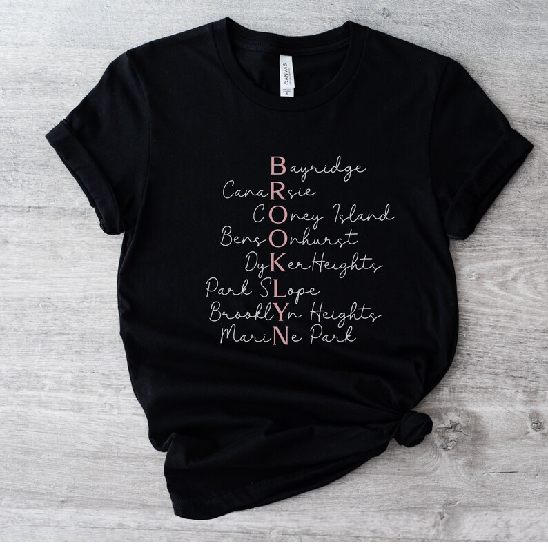 May include: Black t-shirt with the word "BROOKLYN" spelled out vertically with the names of different neighborhoods in Brooklyn, New York, written in pink.
