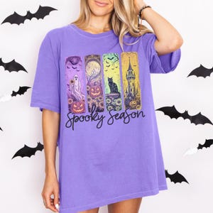 May include: A purple t-shirt with a "Spooky Season" graphic. The graphic features a ghost, jack-o'-lanterns, a black cat, and a haunted tower. Black bat decorations are in the background.