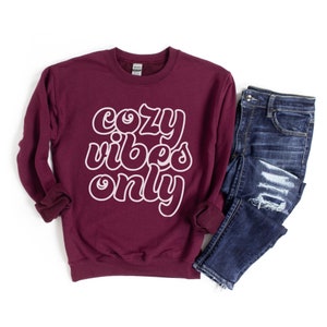 May include: A maroon sweatshirt with the text "cozy vibes only" in white outlined letters. The sweatshirt is shown with a pair of blue jeans.