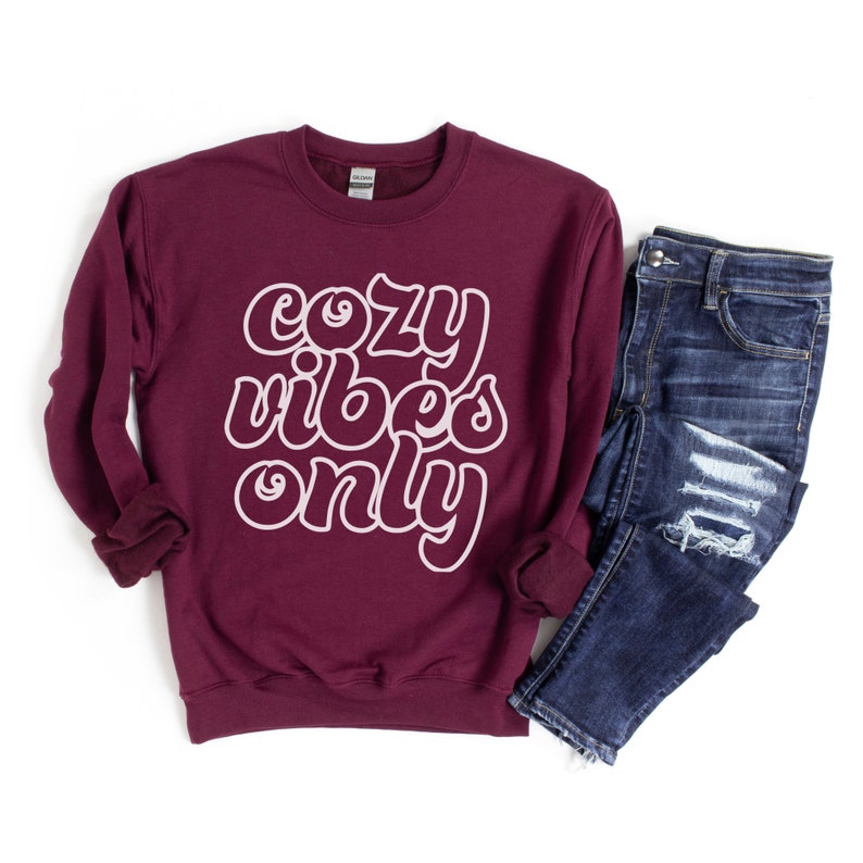 May include: A maroon sweatshirt with the text "cozy vibes only" in white outlined letters. The sweatshirt is shown with a pair of blue jeans.