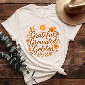 Pu&ograve; includere: T-shirt color crema con le parole "Grateful, Grounded, Golden" in un design circolare. Il design include una luna crescente, un sole ed elementi floreali nei toni dell'arancione e del giallo. &Egrave; visibile un cappello color beige.