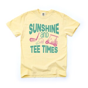 以下が含まれることがあります： 薄い黄色のTシャツ。テキスト「SUNSHINE and TEE TIMES」は、ティールとピンクで表示されています。デザインには、ゴルフクラブ、ゴルフボール、ゴルフカートが含まれています。このシャツは半袖、クルーネックです。