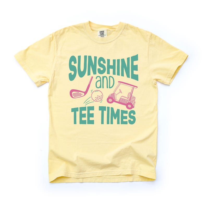 以下が含まれることがあります： 薄い黄色のTシャツ。テキスト「SUNSHINE and TEE TIMES」は、ティールとピンクで表示されています。デザインには、ゴルフクラブ、ゴルフボール、ゴルフカートが含まれています。このシャツは半袖、クルーネックです。