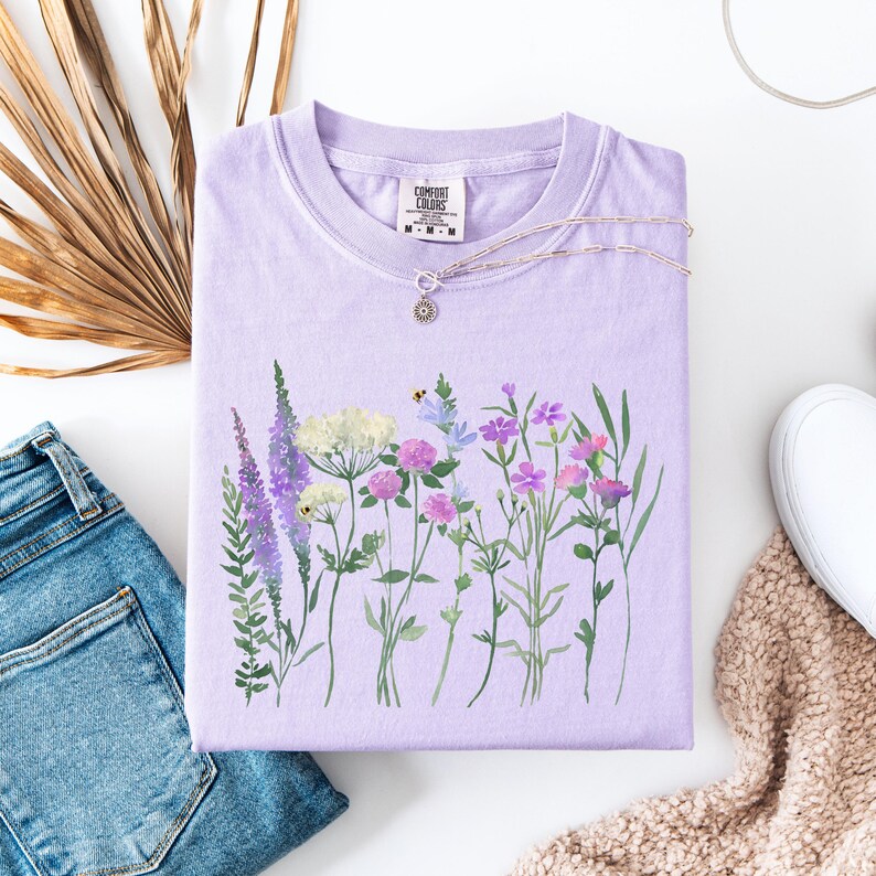 May include: A lavender Comfort Colors t-shirt with a watercolor floral design. The design features various wildflowers in shades of purple, pink, white, and green. A silver necklace with a small pendant rests on the shirt. The shirt is folded, with a pair of blue jeans and a sneaker visible.