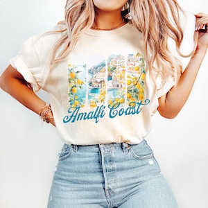 Pu&ograve; includere: Una maglietta bianca con un'immagine ad acquerello della Costiera Amalfitana, in Italia, con alberi di limone e il testo "Amalfi Coast" in blu.