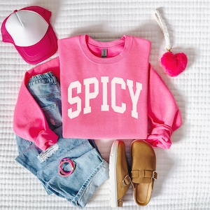 K&ouml;nnte beinhalten: Ein rosa Sweatshirt mit dem Wort "SPICY" in wei&szlig;en Blockbuchstaben. Das Sweatshirt liegt auf einer wei&szlig;en Oberfl&auml;che mit einer blauen Jeans, braunen Clogs, einer rosa Trucker-M&uuml;tze und einem rosa flauschigen Herzen.