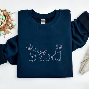 May include: A navy blue sweatshirt featuring a graphic of three bunnies in a row. The bunnies are white with pink noses and inner ears.
