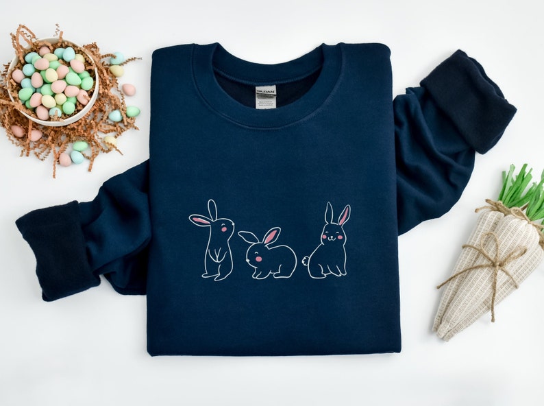 May include: A navy blue sweatshirt featuring a graphic of three bunnies in a row. The bunnies are white with pink noses and inner ears.