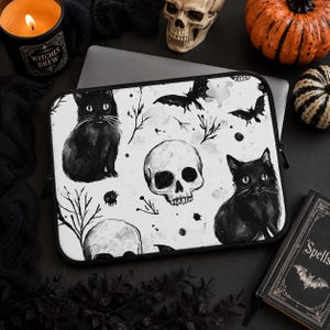 Può includere: Una custodia rettangolare per computer portatile con sfondo bianco e un motivo a inchiostro nero raffigurante due gatti neri seduti, due teschi umani, pipistrelli in volo e rami d'albero spogli. La custodia ha una cerniera nera.