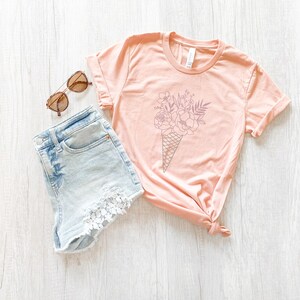 May include: A light pink t-shirt with a graphic of a bouquet of flowers in an ice cream cone. The shirt is paired with light blue denim shorts with lace trim.